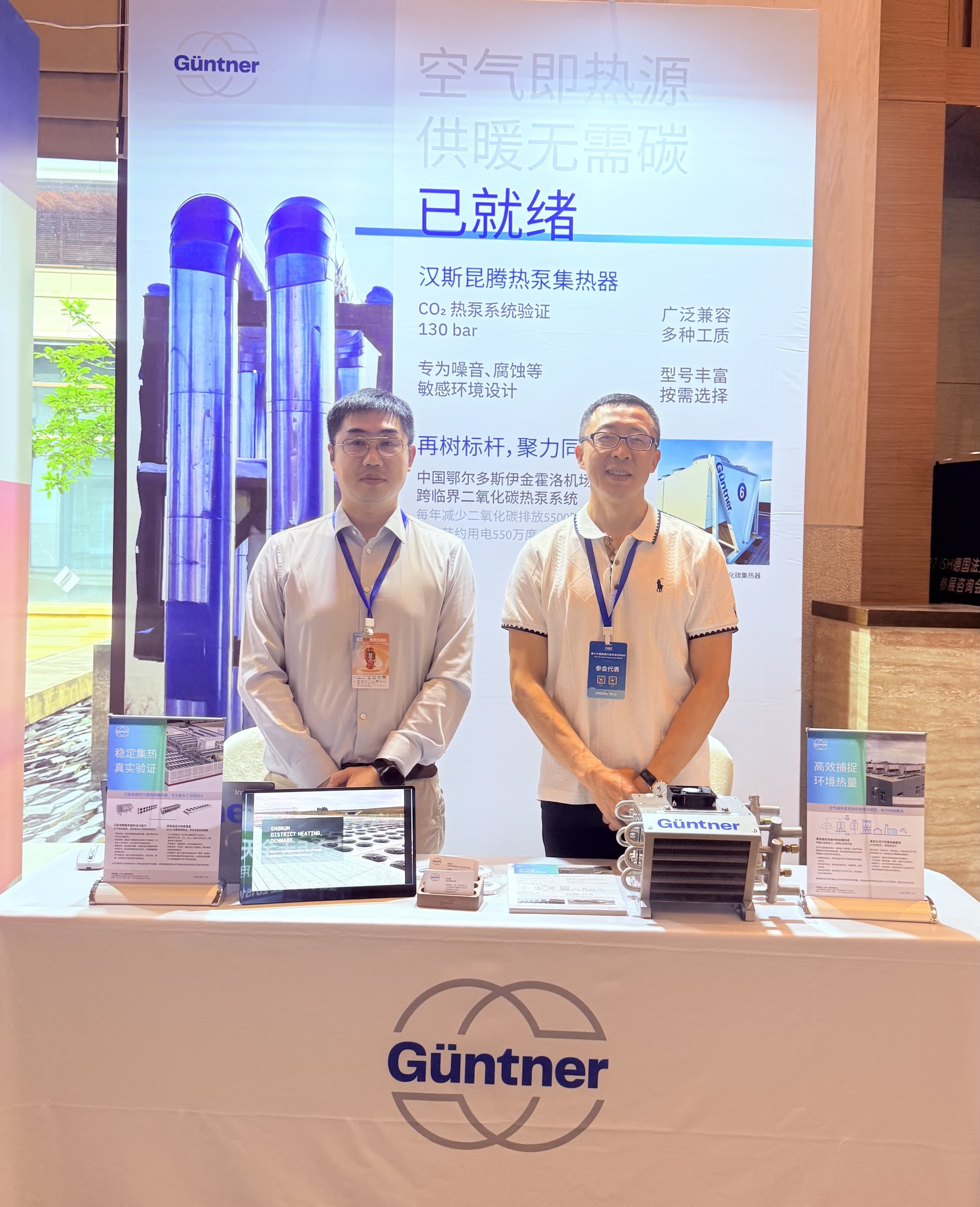 Two men with lanyards stand behind a display booth featuring a large banner with Chinese text and purple-tipped cooling towers; Güntner logo on table front.