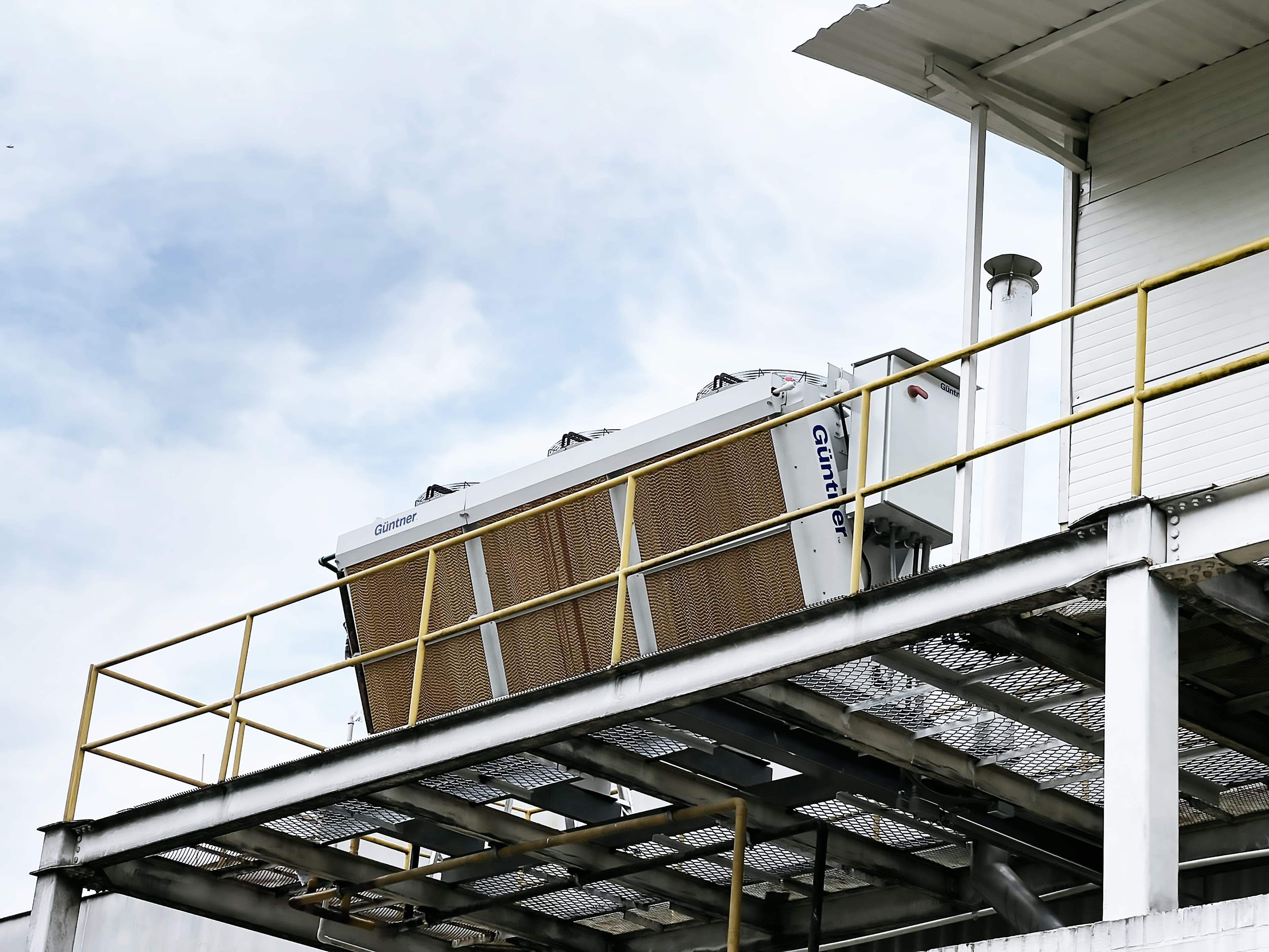 Équipement industriel sur une plateforme métallique avec garde-corps jaune; unité de refroidissement blanche marquée Guntner, ciel bleu en arrière-plan.