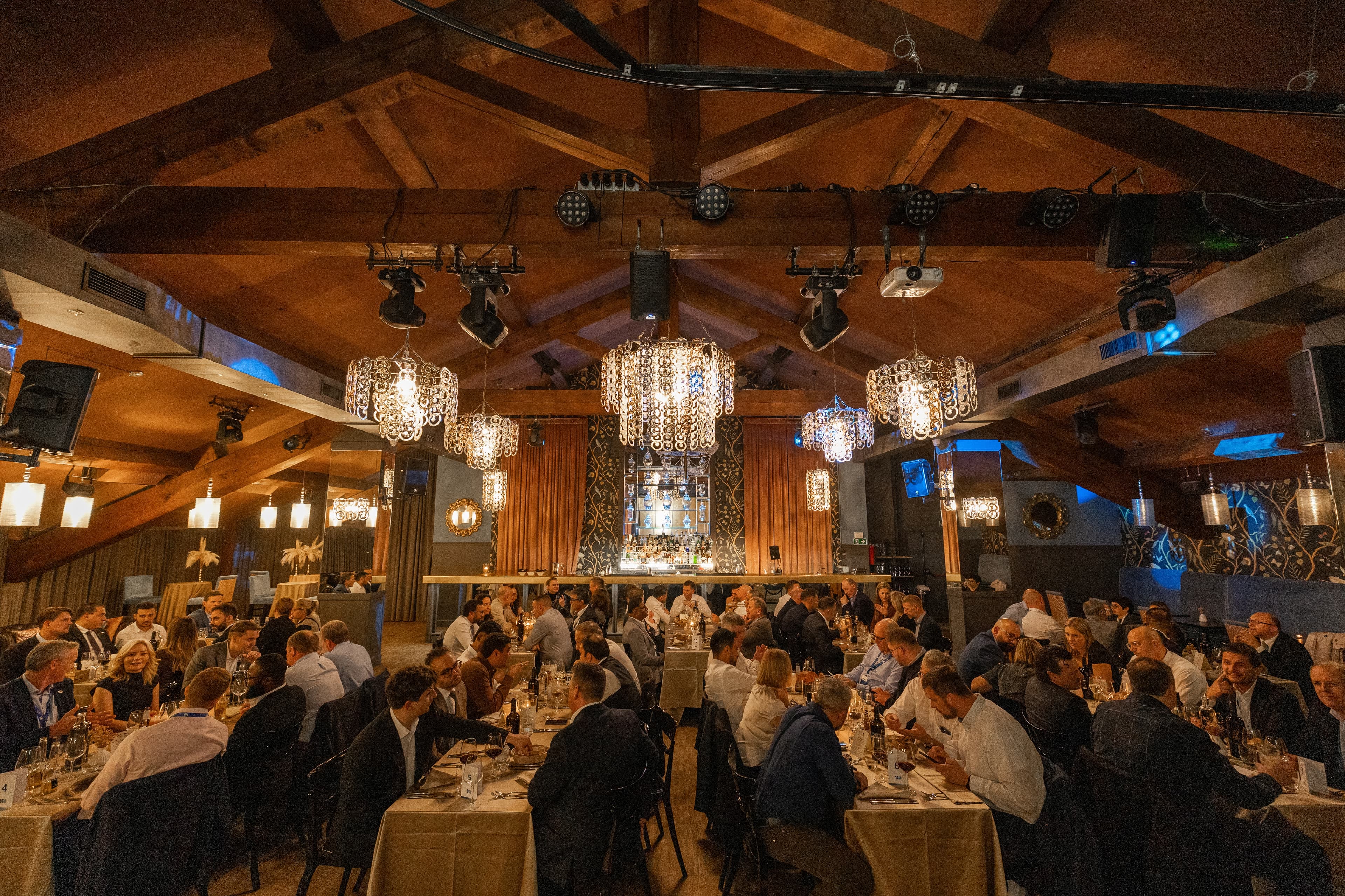 Banquet hall with exposed wooden beams, ornate chandeliers and long rows of tables filled with formally dressed guests dining under warm lighting.