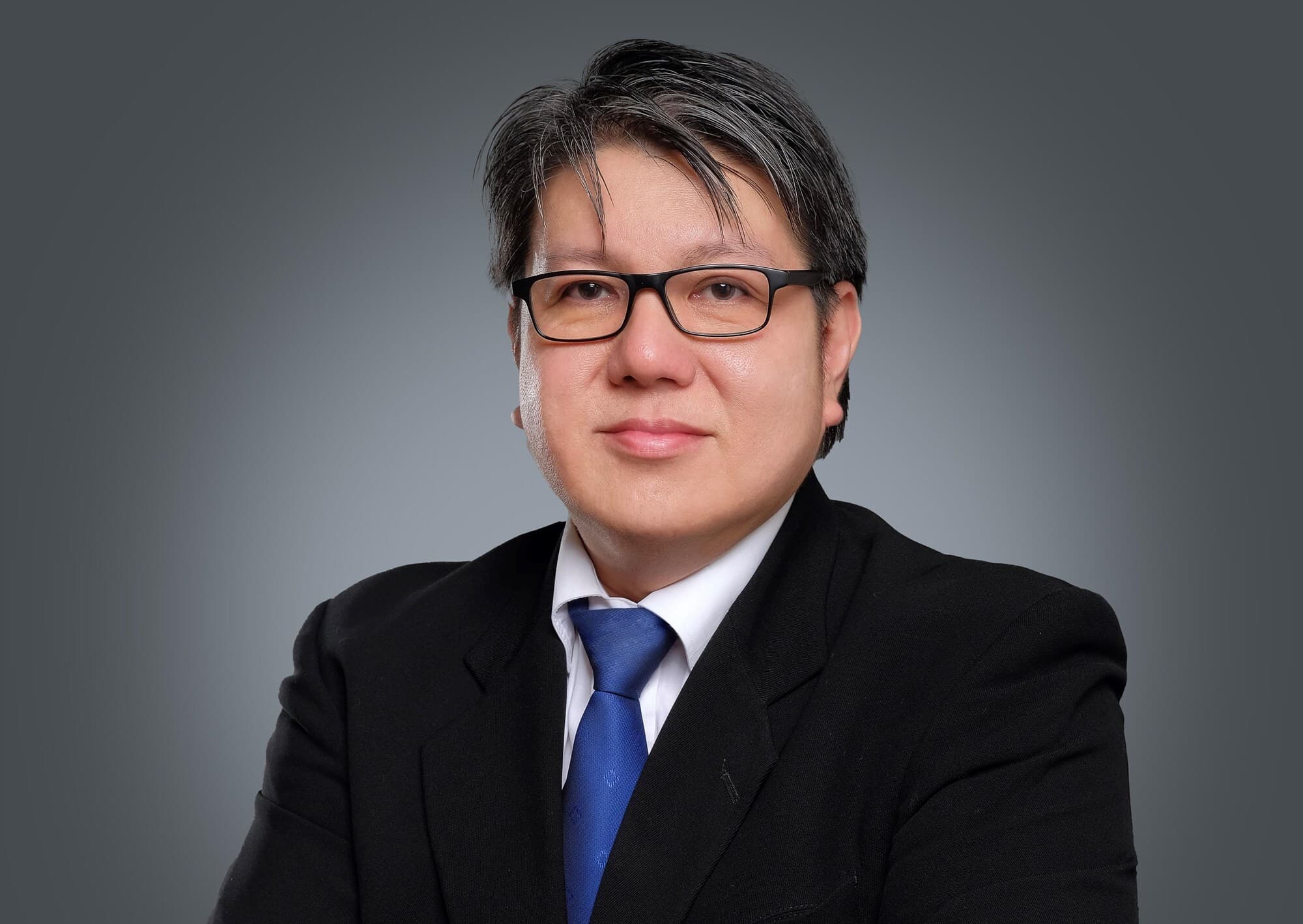 Head-and-shoulders portrait of an individual with short dark hair and glasses, wearing a black suit, white shirt, and blue tie, against a gray gradient background.