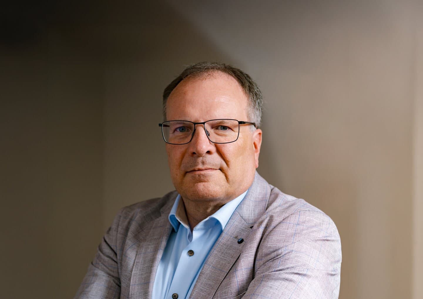 Person with short grey hair and glasses, wearing a light blue shirt and a light checkered blazer, looking at the camera against a neutral background.