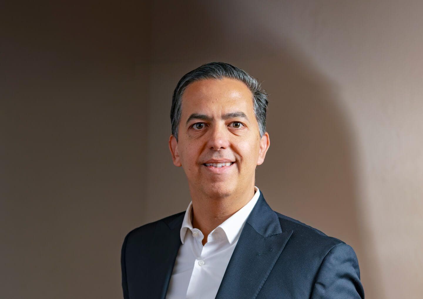 Portrait of a person with short, graying hair wearing a dark blazer and white shirt, smiling against a neutral beige background.