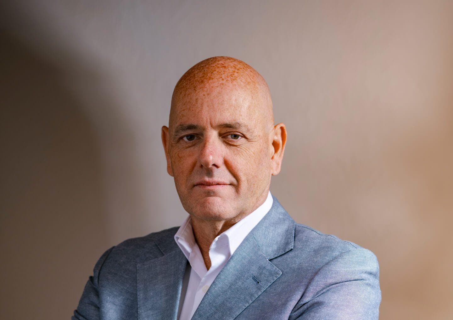 Head-and-shoulders portrait of a bald person with freckles, wearing a light blue blazer and white shirt, against a neutral gradient background.