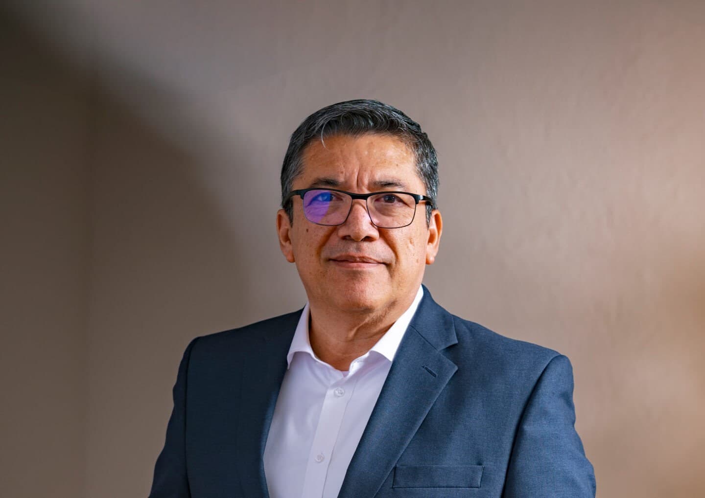 Middle-aged person with short dark hair and glasses, wearing a navy blazer and white shirt, against a plain beige background.