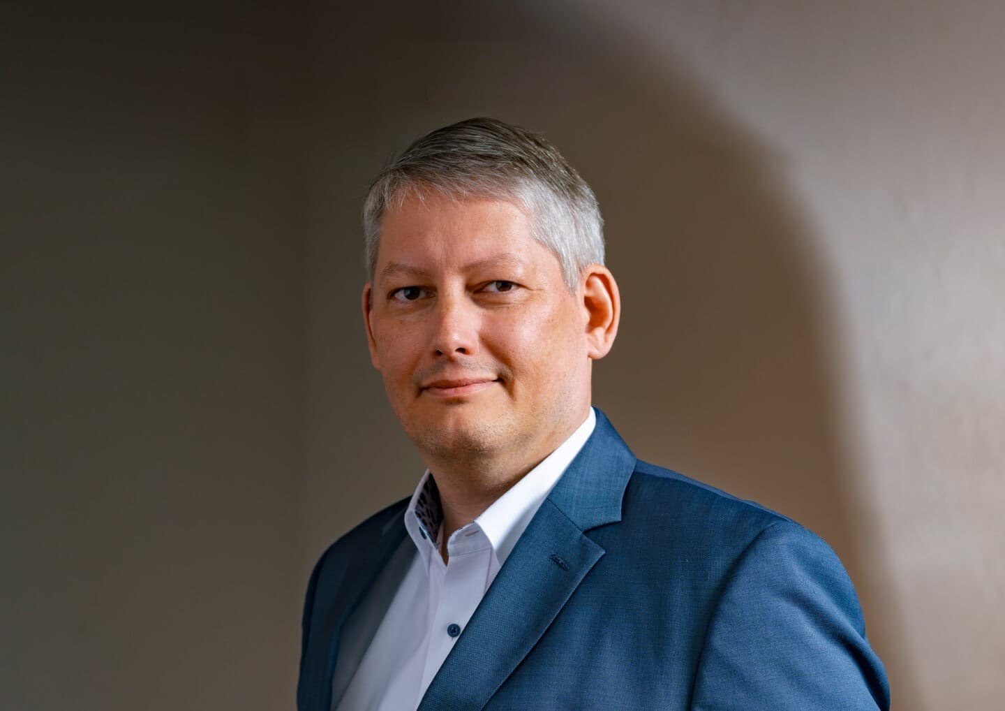 Portrait of a person with short gray hair, wearing a blue suit and white shirt, looking toward the camera with a subtle smile, against a neutral backdrop.