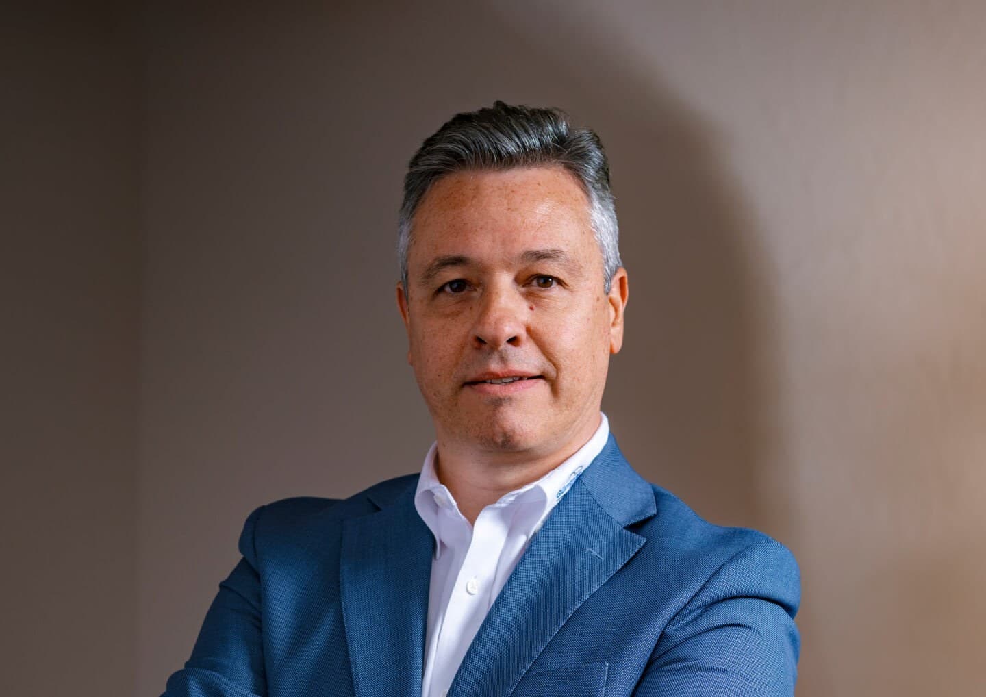 A person with short gray hair in a blue blazer and white shirt stands against a neutral beige background, looking at the camera with a subtle smile.