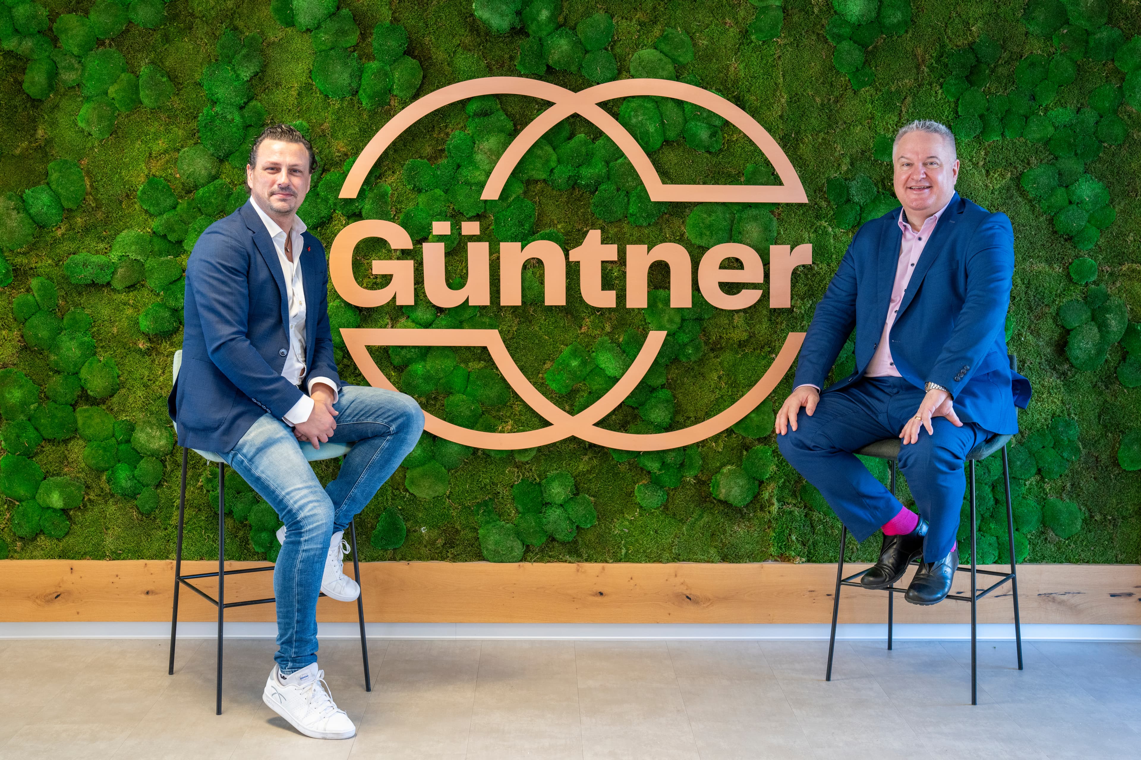 Two people sit on high stools in front of a green moss wall with a large orange Güntner logo and overlapping arches.