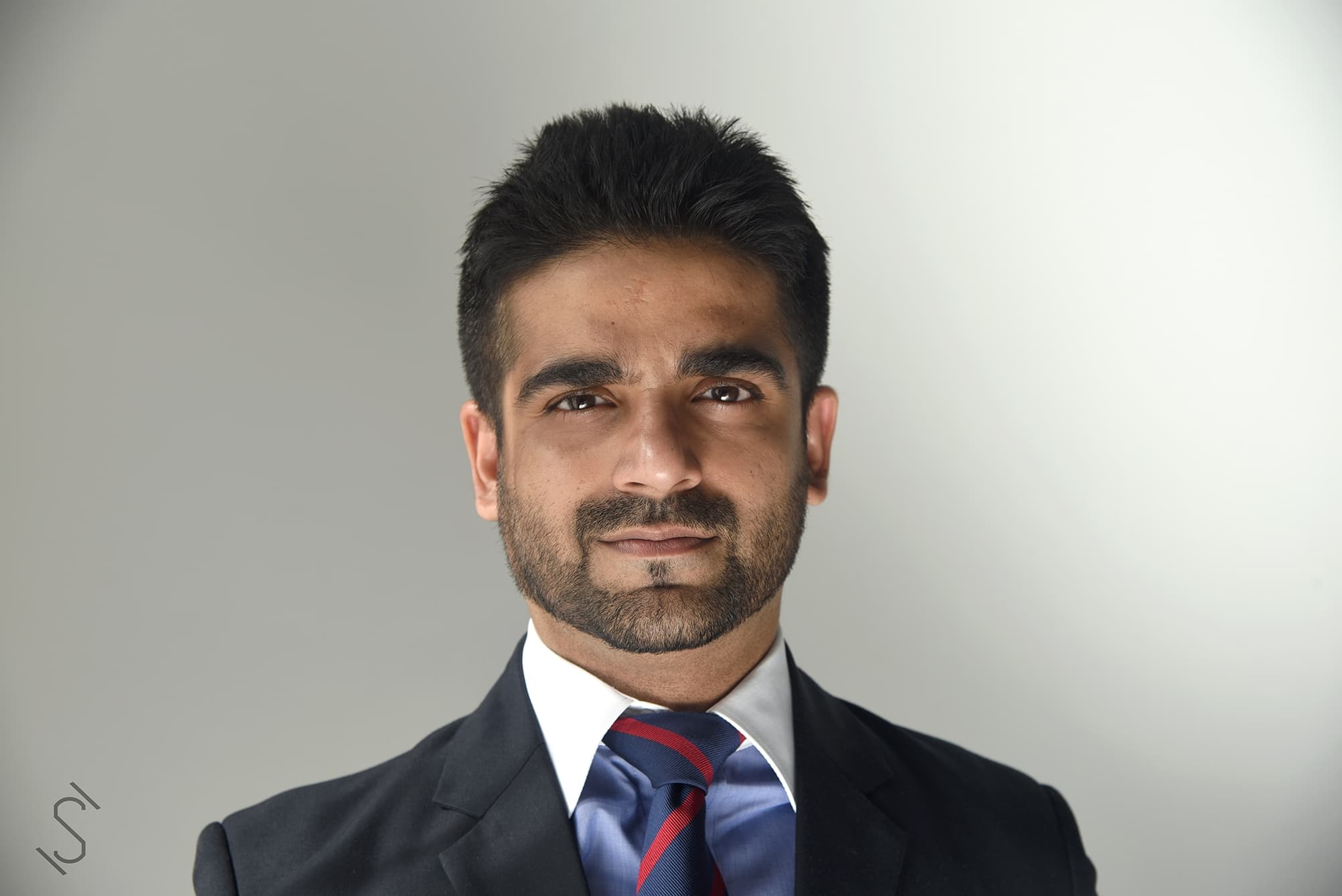 Portrait of a person with short dark hair and a trimmed beard, in a dark suit, white shirt, and blue-red striped tie, against a neutral gray background.