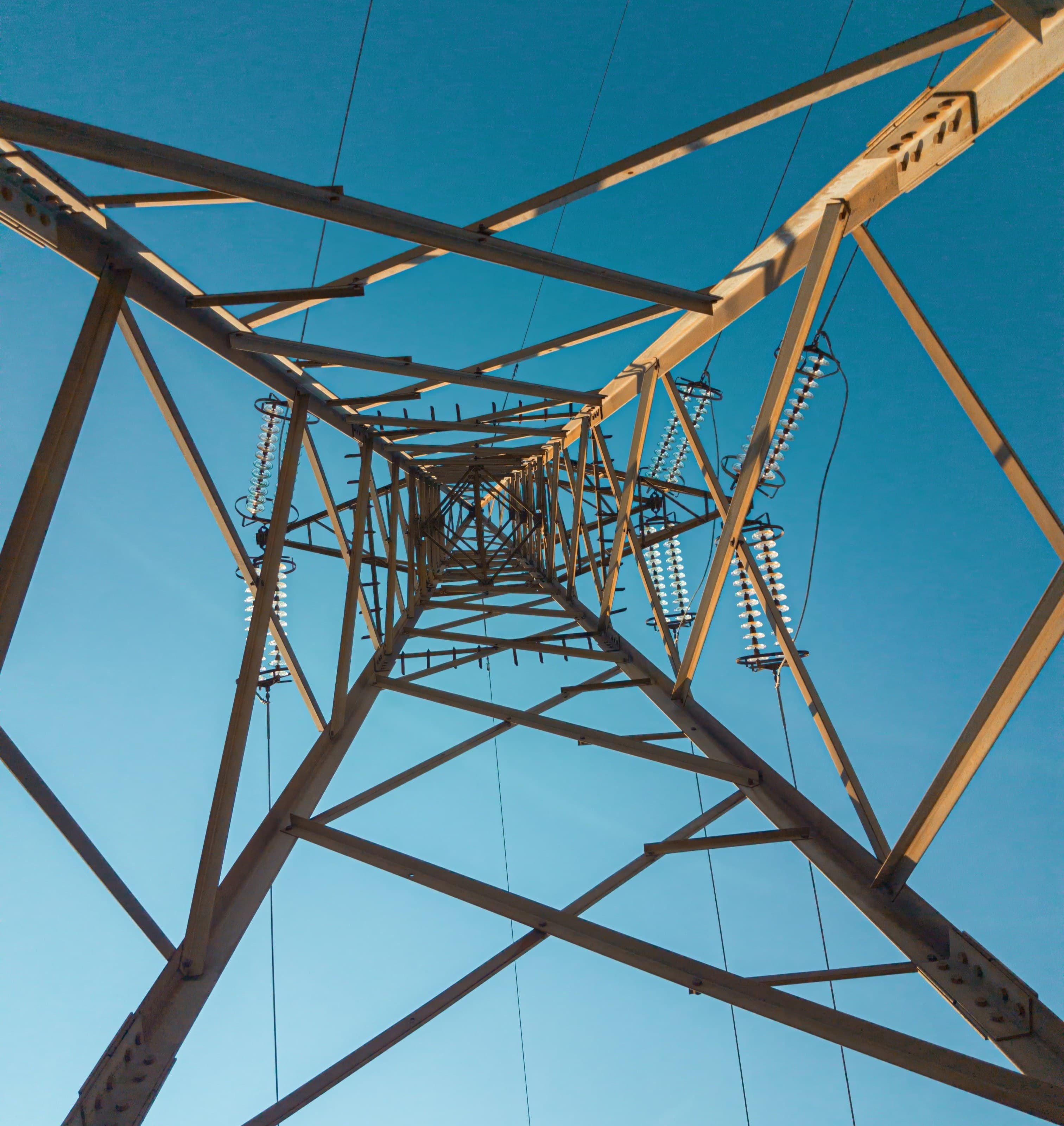 View from below of a steel lattice power transmission tower, with insulators along the arms, converging towards the apex against a clear blue sky.