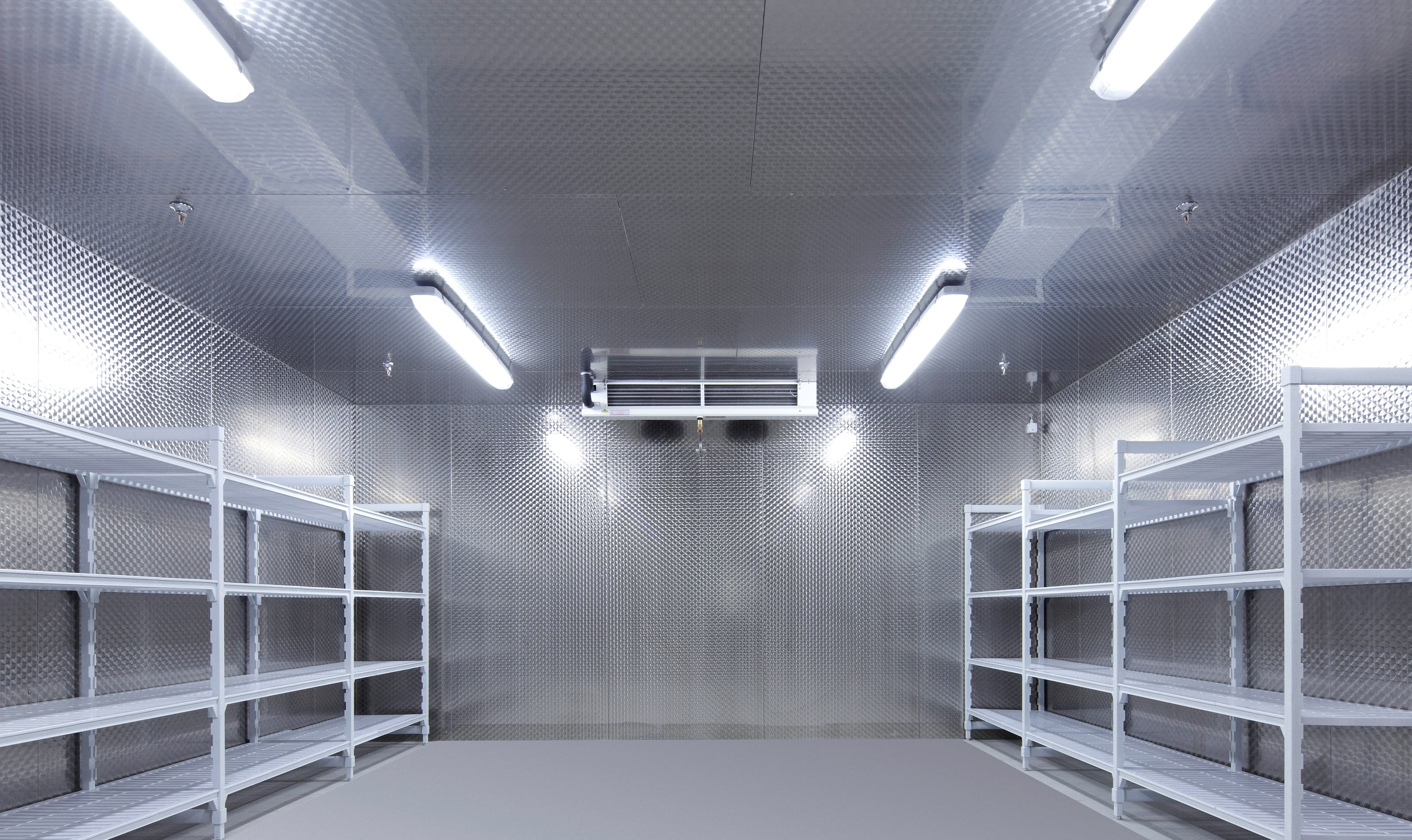 Empty metal storage room with diamond-plate walls, white metal shelving on both sides, and a ceiling air conditioner under bright fluorescent lights.