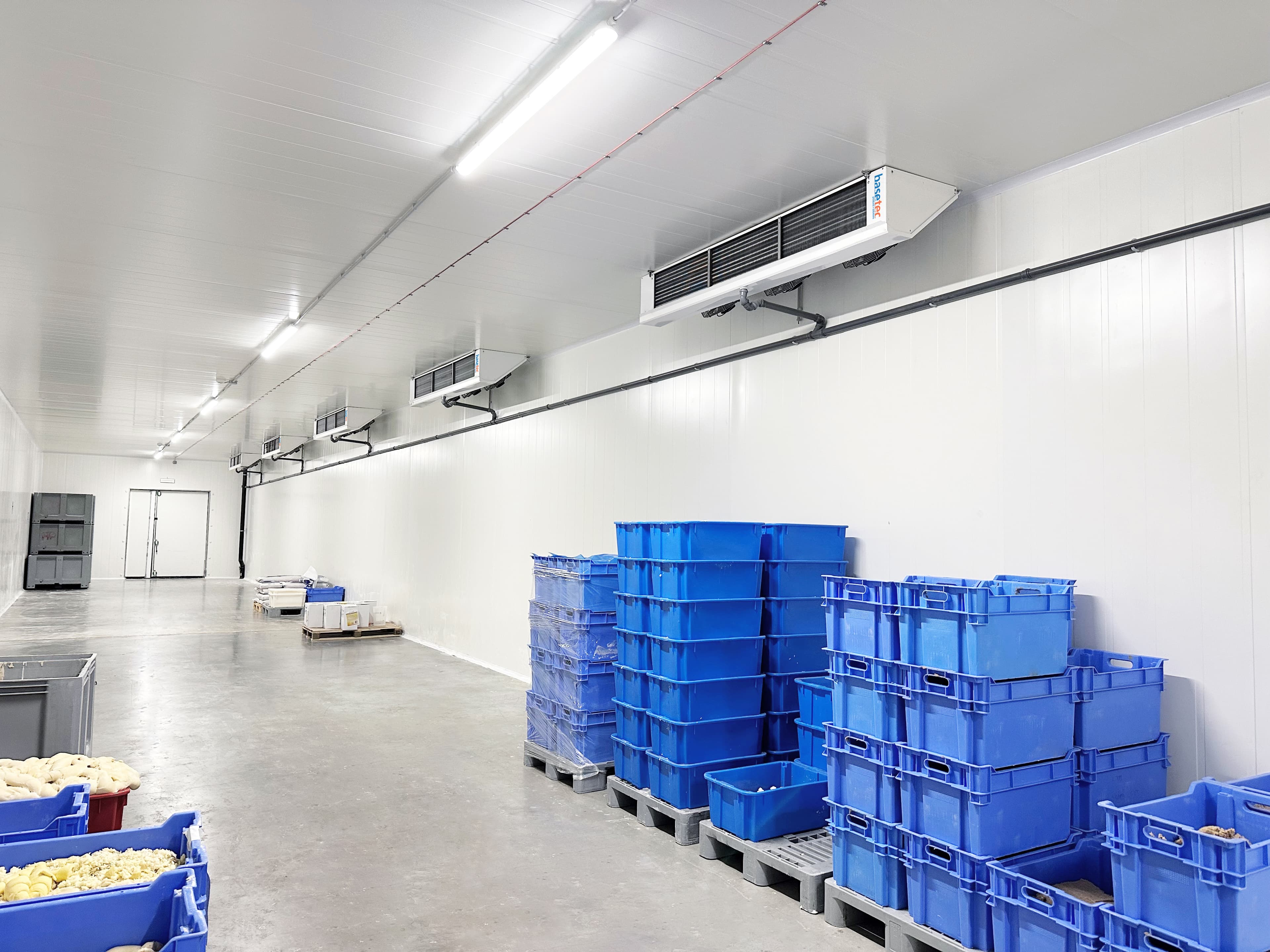 Clean industrial storage room with white walls and ceiling, bright overhead lights, and blue plastic crates stacked on pallets along the right wall.
