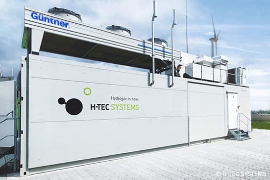 A large white modular industrial unit on a concrete pad, bearing Güttner and H-TEC SYSTEMS logos, with rooftop vents and a distant wind turbine against a blue sky.