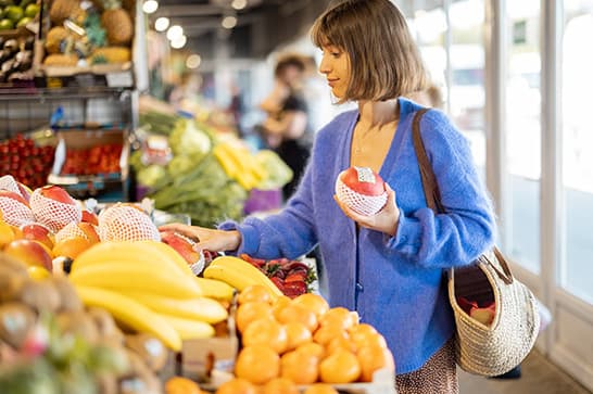 A person with short brown hair in a blue cardigan browses fruit at a grocery store, holding a red apple and a woven bag among bananas and oranges.