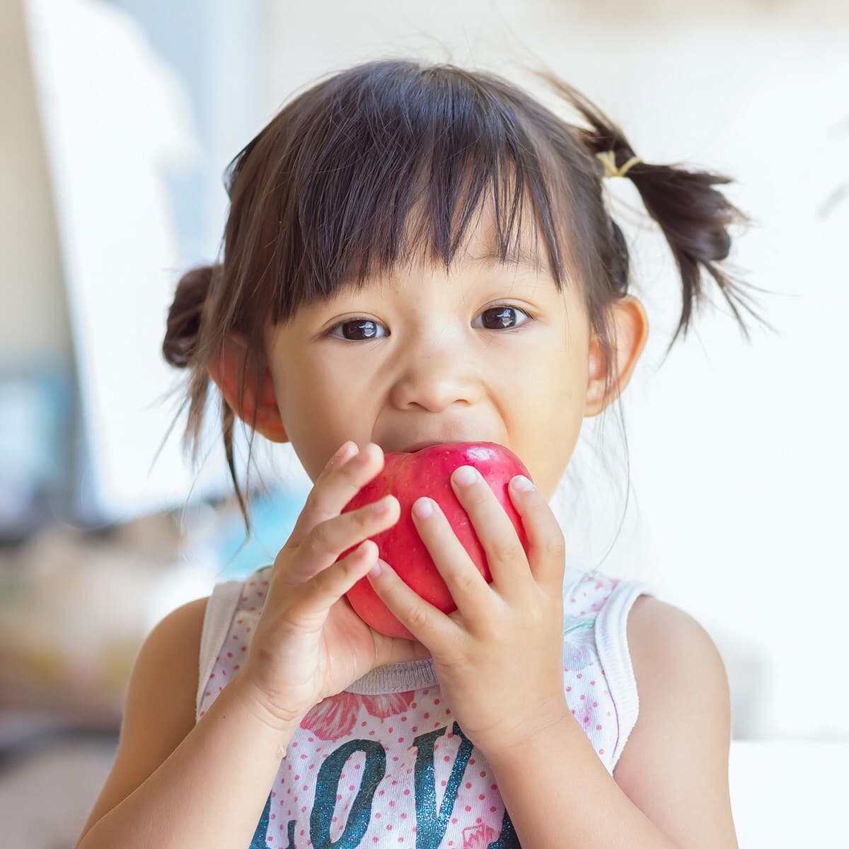 A young child with dark twin ponytails bites into a red apple, looking at the camera.