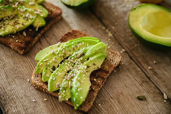 Toast slices topped with avocado slices, sesame seeds, and a light sprinkle on a rustic wooden surface, with a halved avocado nearby.