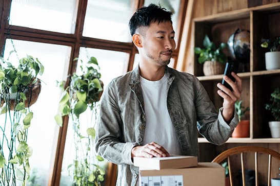 Person with short dark hair in a light jacket and white shirt stands by a plant-filled window, looking at a smartphone while handling a box.