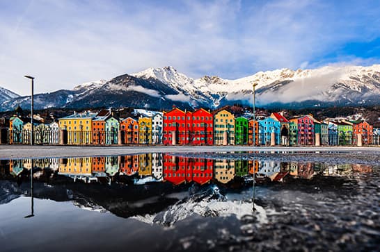 Row of colorful houses along a calm waterfront with snow-capped mountains in the background; vibrant reflections mirror on the still water.