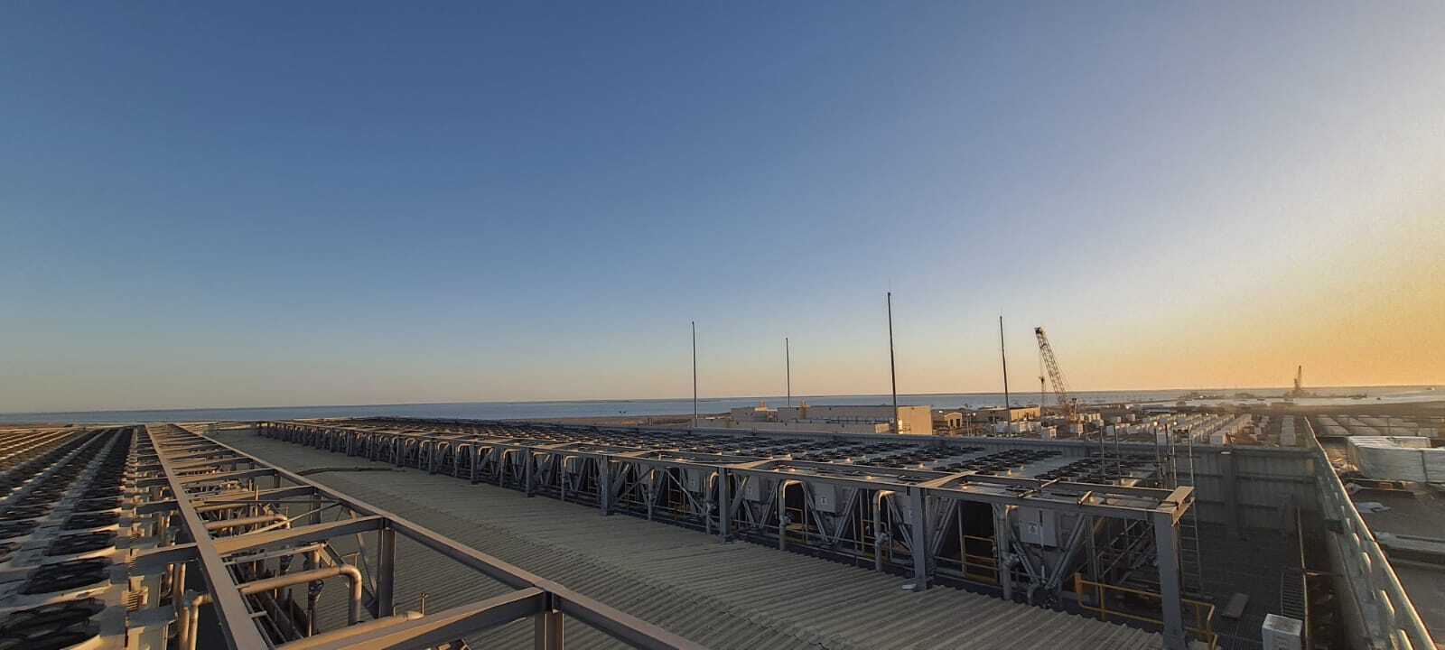 Coastal industrial site with rows of metal framework and walkways, a crane in the distance, calm sea and warm sunset sky on the horizon.