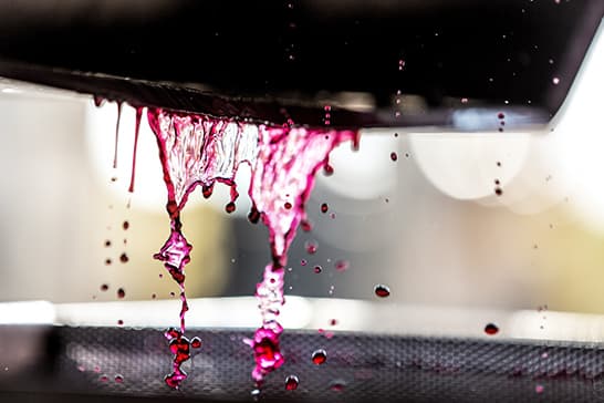 Close-up of neon pink liquid dripping from a dark surface onto a perforated tray, with scattered droplets and a softly blurred background.