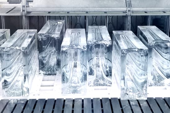 Clear rectangular ice blocks resting on a white tray in an industrial ice production area, with metal shelving and a slatted floor in the background.