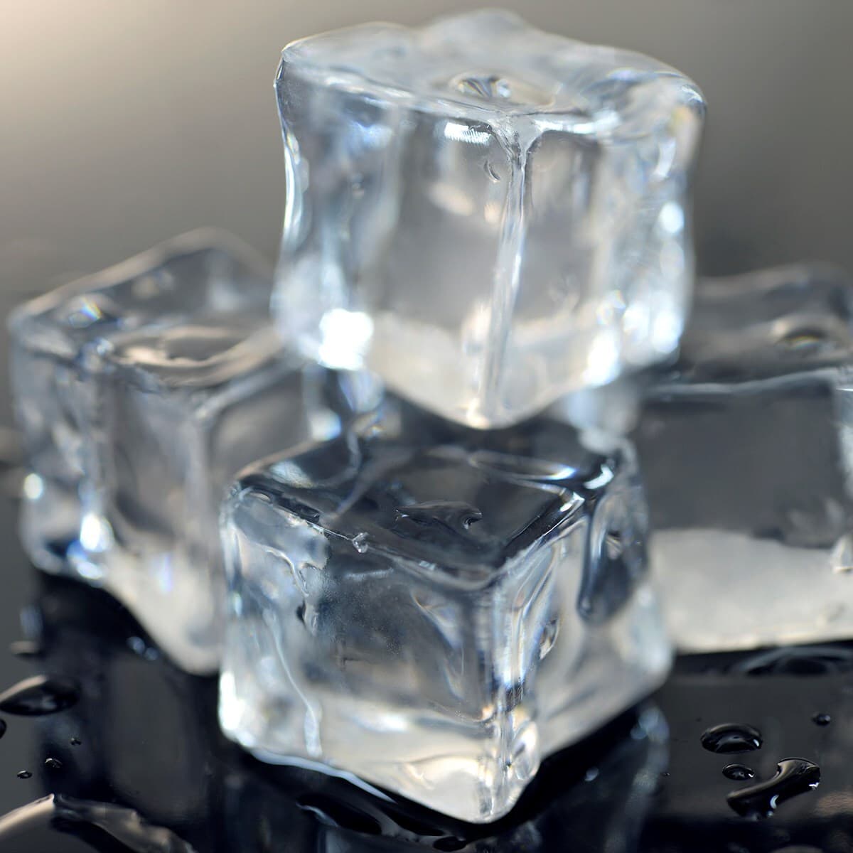 Close-up of clear ice cubes stacked on a dark glossy surface, with reflections and small water droplets.