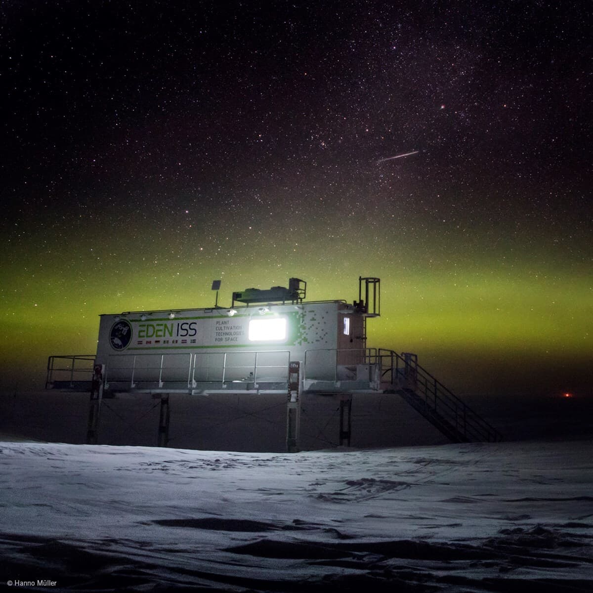Nighttime view of a raised research module labeled EDEN ISS on stilts above a snow-covered landscape, beneath a green aurora and a starry sky.
