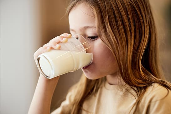 A person with long light brown hair in a beige top drinks milk from a clear glass, held to the lips in a softly lit indoor setting.