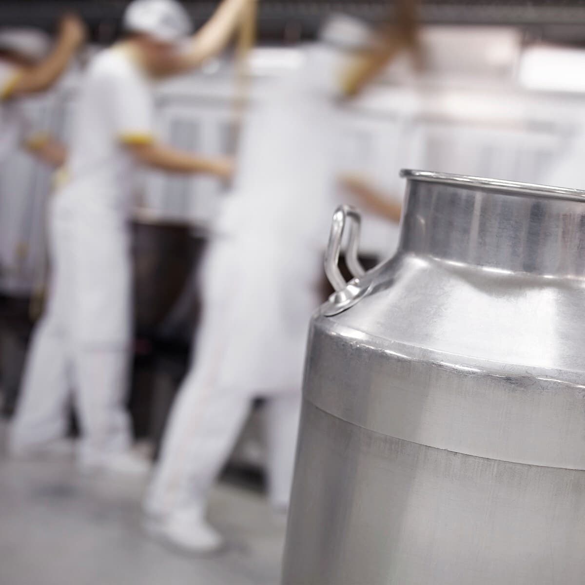 Close-up of a shiny metal milk can in the foreground with blurred workers in white outfits in a dairy processing facility.
