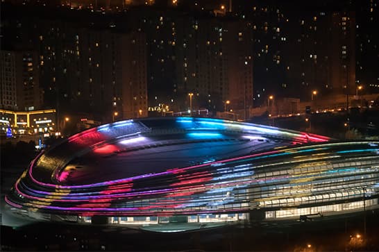 Nighttime aerial view of an oval stadium with bright multicolor LED stripes on its curved roof, set against a glowing city skyline.