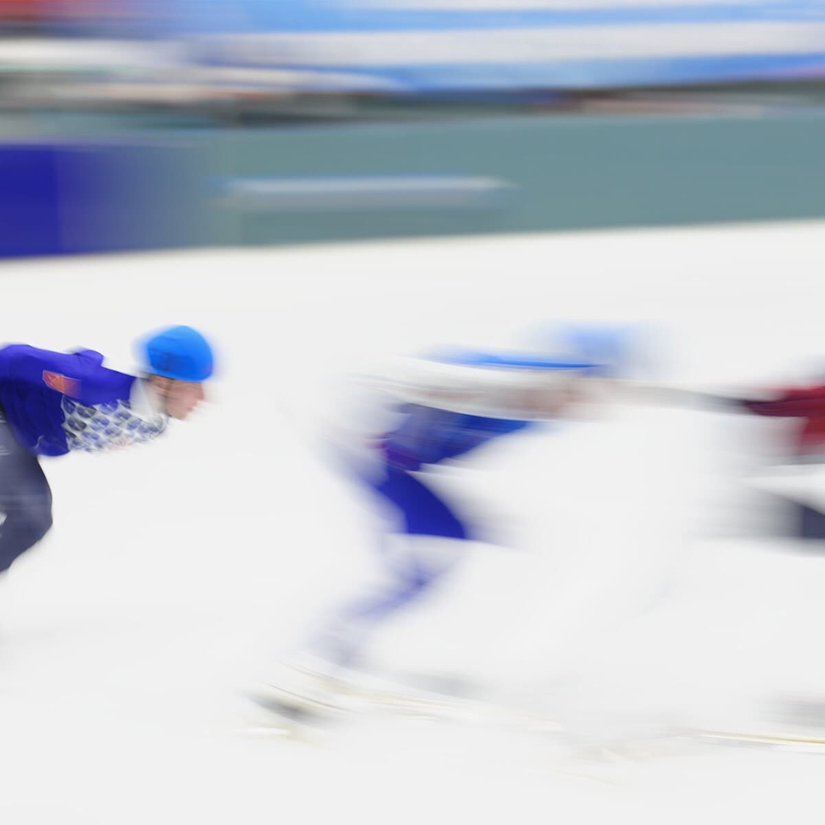 Three skaters in blue helmets and outfits glide quickly across a bright ice rink, captured with motion blur against a blue and white background.