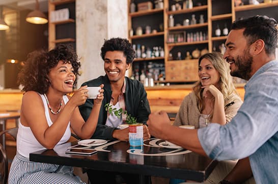 Four friends sit at a cafe table, smiling and chatting as one lifts a cup; warm lighting and shelves full of bottles in the background.