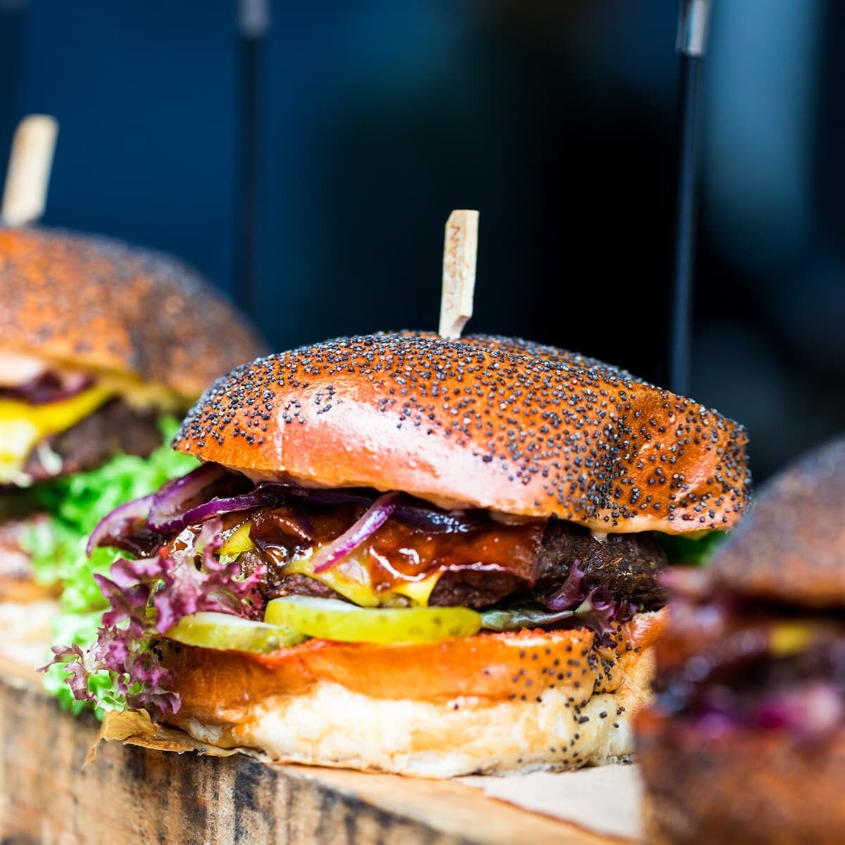 Close-up of a sesame-seed burger with lettuce, pickles, red onions, cheese, and sauce on a toasted bun, skewered with a wooden toothpick.