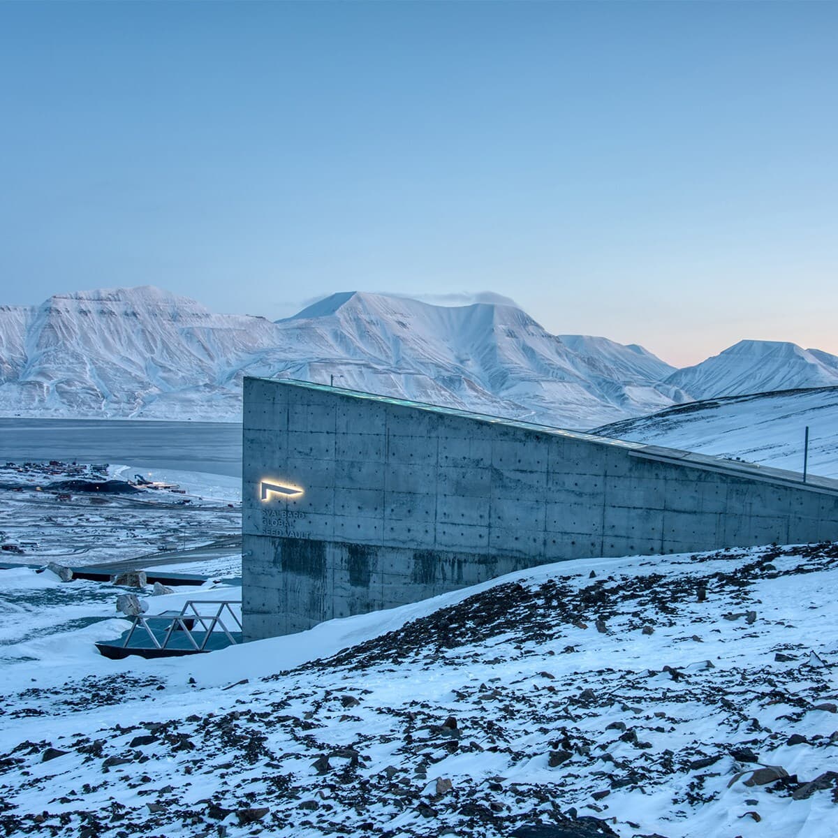 Snowy Arctic scene with a slanted concrete building in the foreground, the Svalbard Global Seed Vault, and blue, snow-covered mountains in distance.