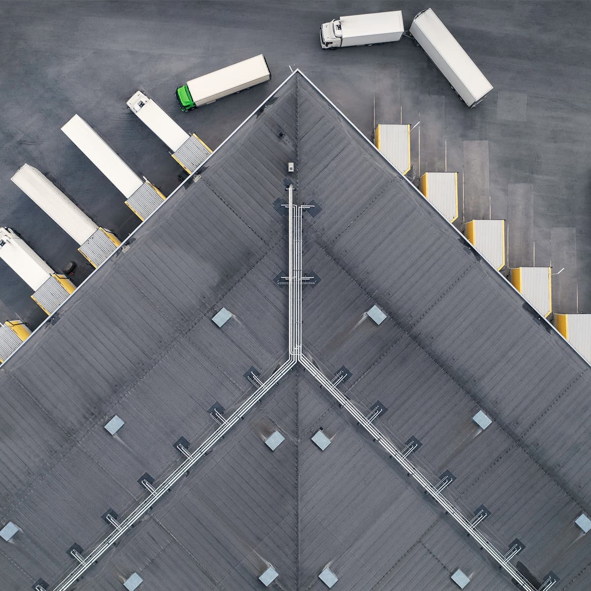 Aerial view of a large gray warehouse with a triangular roof; loading docks line the sides with several white trailers and a green-cab truck.