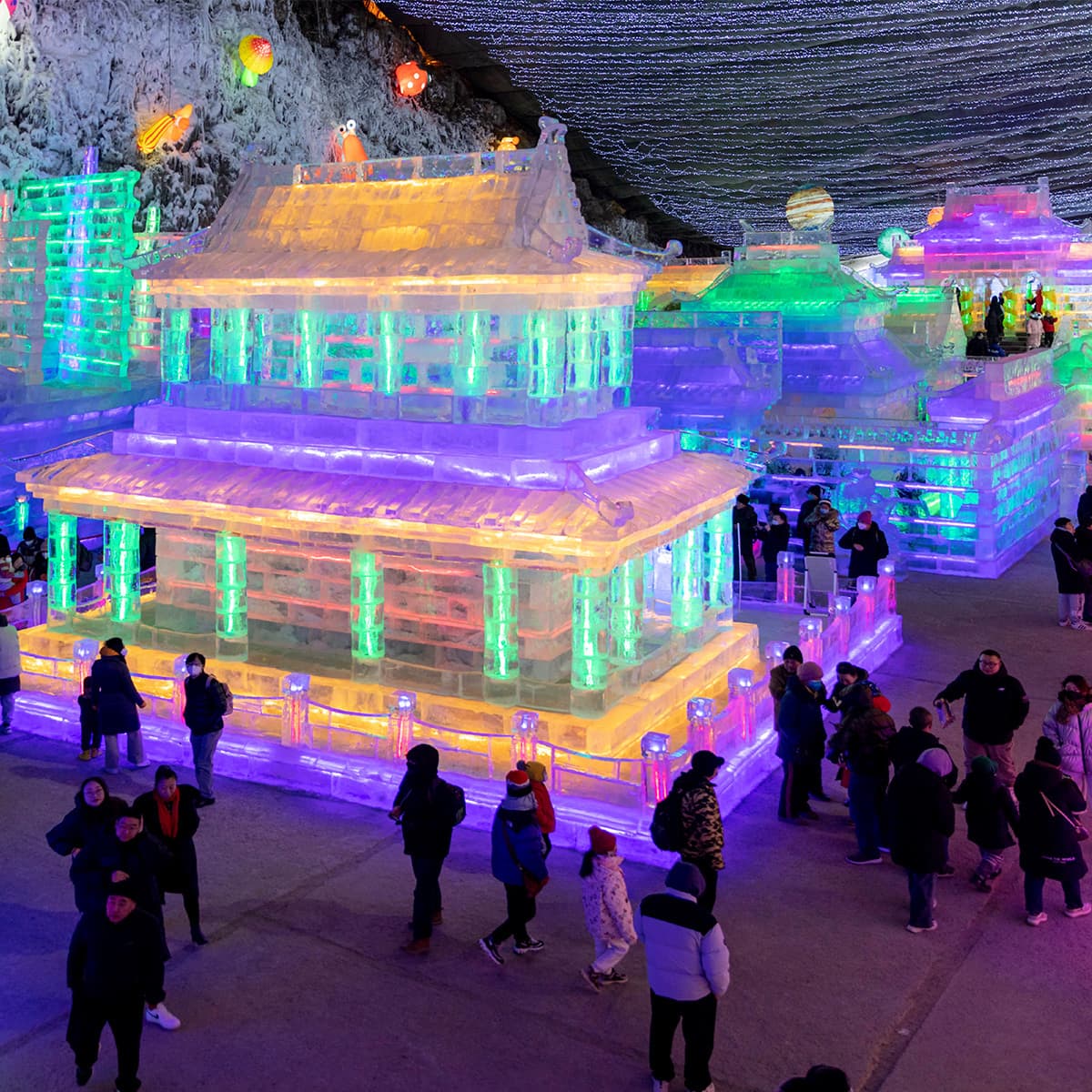 Colorful illuminated ice structures forming a pagoda-style village at night, with visitors in winter coats strolling among glowing blocks and rails.