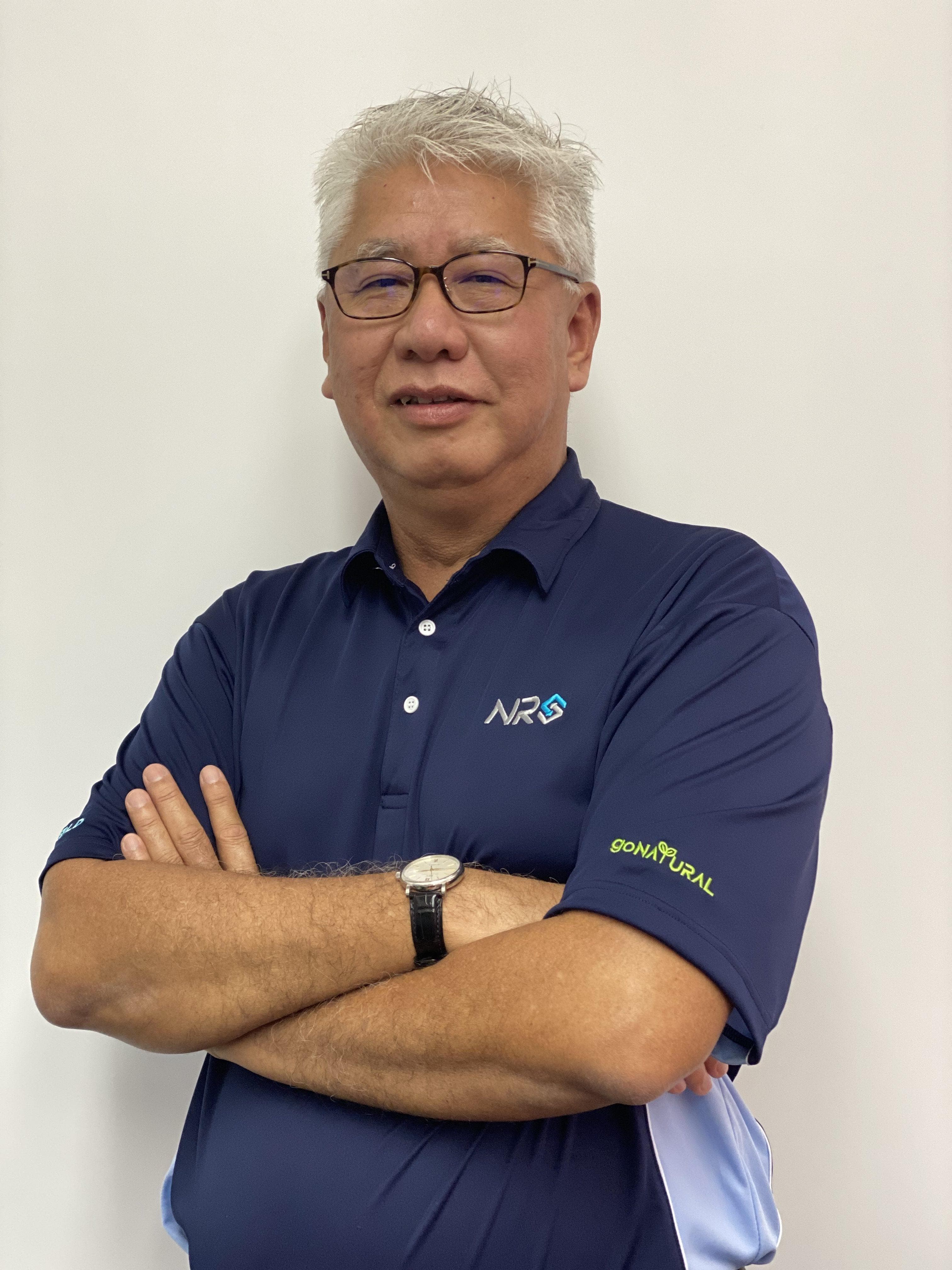 Portrait of an adult with short gray hair and glasses, wearing a navy polo with logos, arms crossed, against a plain light background.