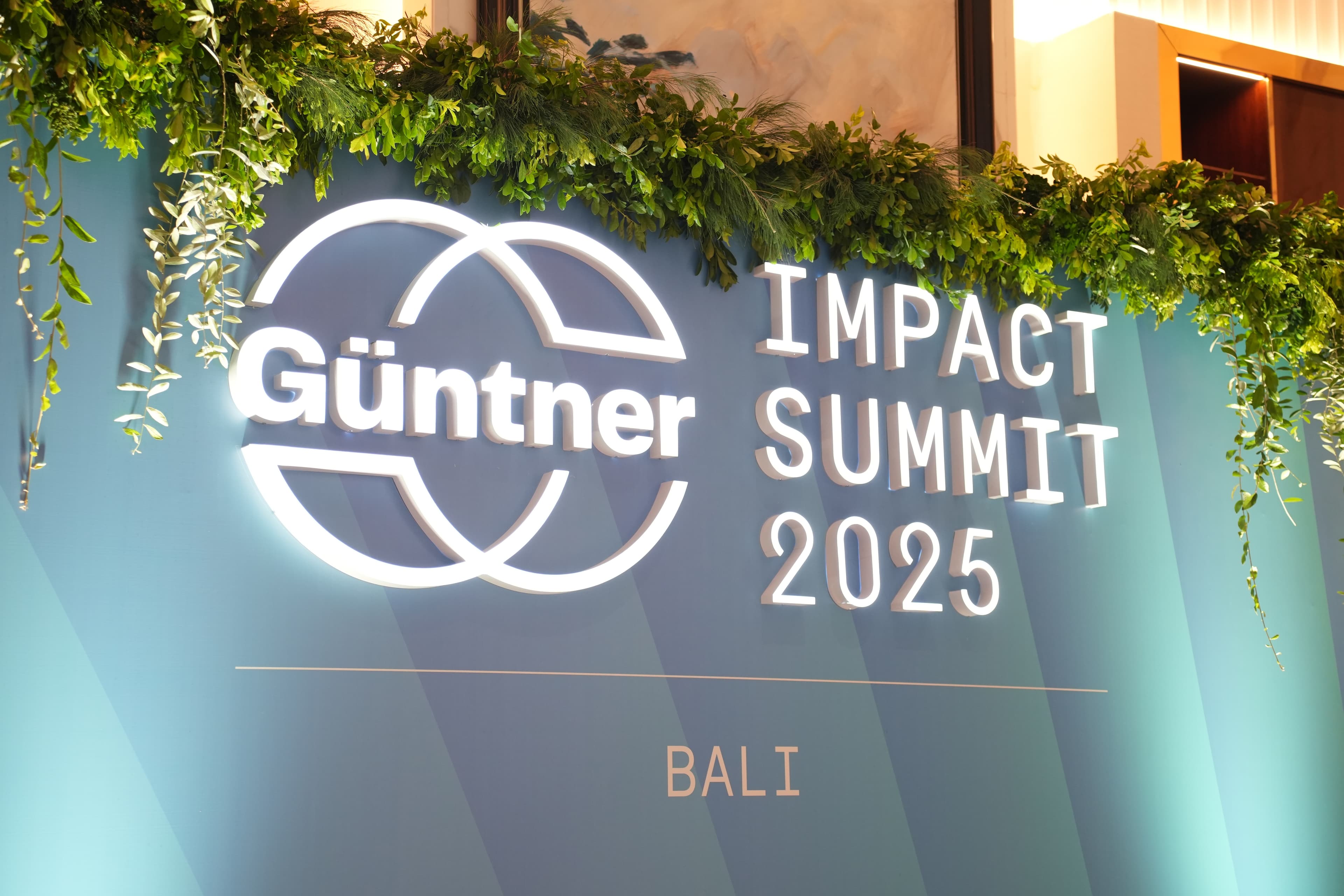 Teal gradient backdrop for Impact Günttner Summit 2025 Bali, with a white circular logo and bold lettering, framed by lush green plants along the top.
