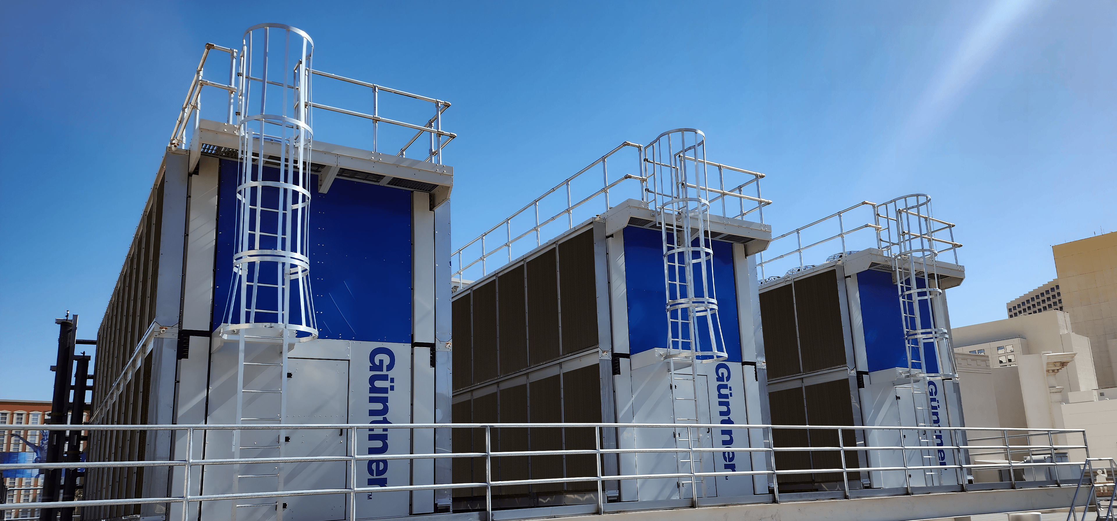 Industrial cooling towers with blue panels, metal staircases, and safety railings on a rooftop under a clear blue sky.