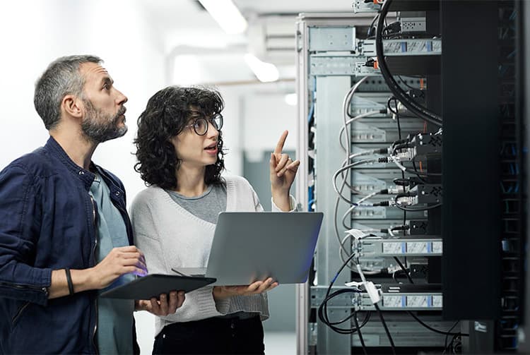 Two people in a data center examine a server rack; one holds a laptop while the other points at equipment amid tangled cables.