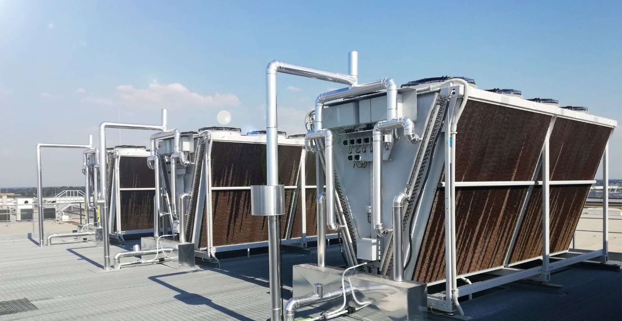 Rooftop industrial cooling system with large brown finned heat exchangers, silver piping, and overhead fans under a clear blue sky.