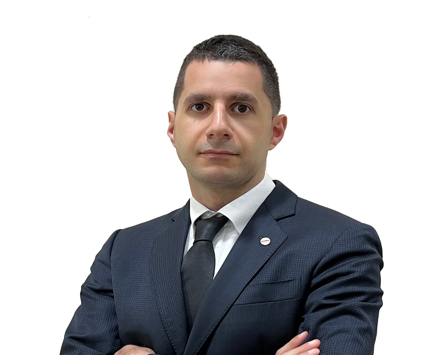 A person in a dark textured suit, white shirt, and black tie, with arms crossed, short dark hair, a neutral expression, against a white background.