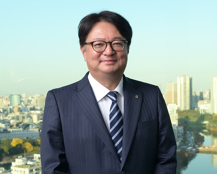 A person in a dark pinstripe suit, white shirt, and striped tie stands outdoors, wearing glasses and smiling, with a city skyline and river behind them.
