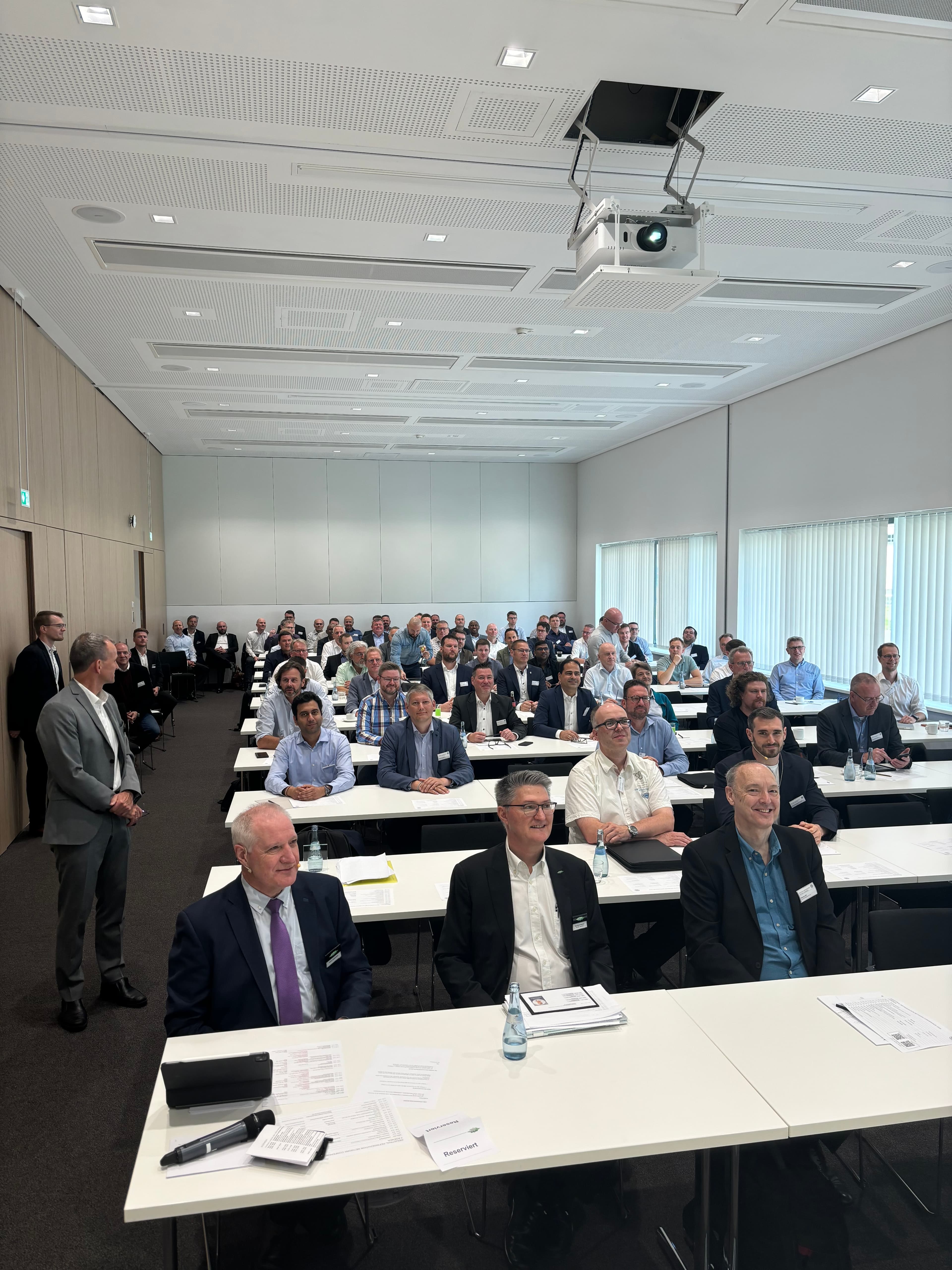 Large conference room filled with attendees in business attire at white tables, facing forward; a ceiling projector hangs above as a person stands to the left.