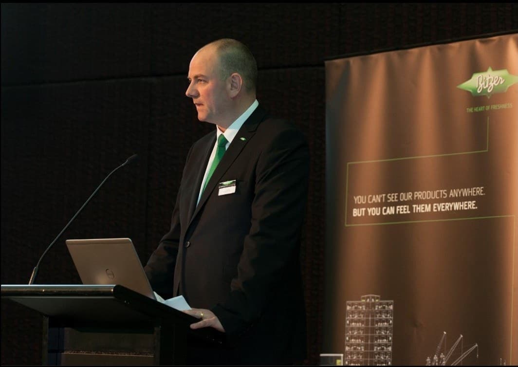 A presenter in a black suit with a green tie stands at a podium with a laptop and microphone, beside a tall branded banner.