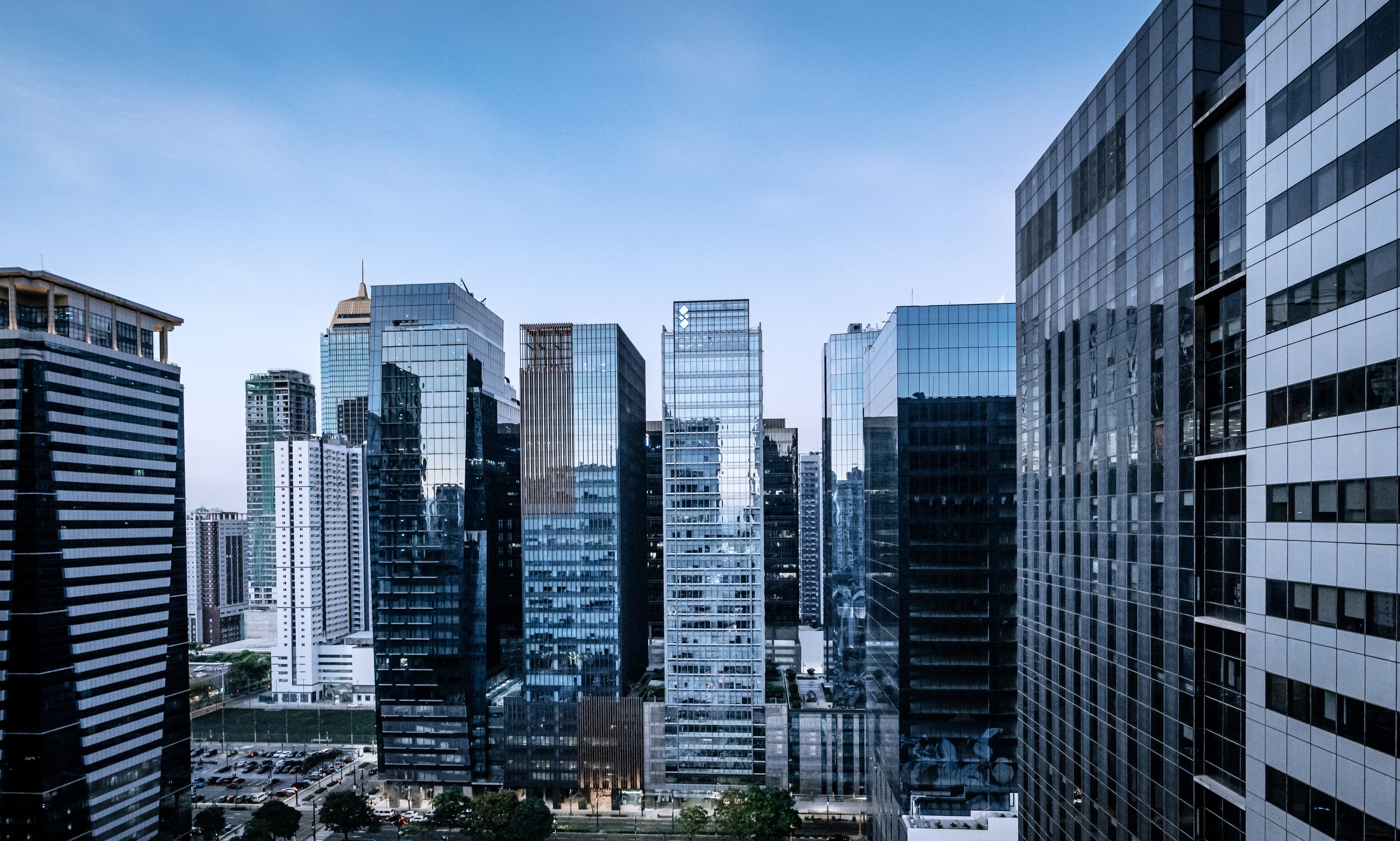City skyline of tall glass skyscrapers, viewed from above, under a clear blue sky; streets and greenery visible at the base.