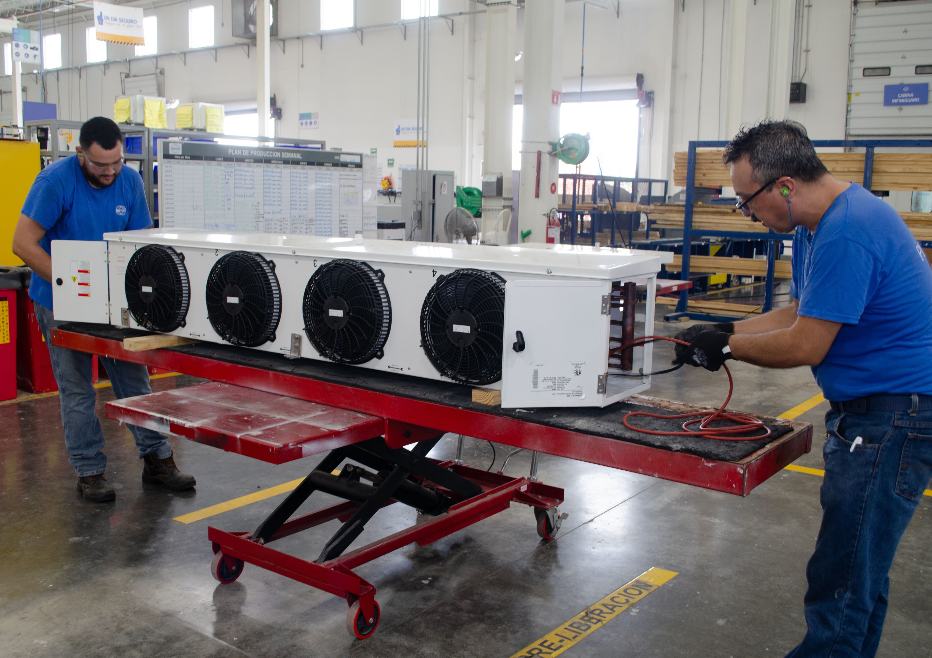 Two workers in blue shirts assemble a white cooling unit with four fans on a red mobile platform in a factory.