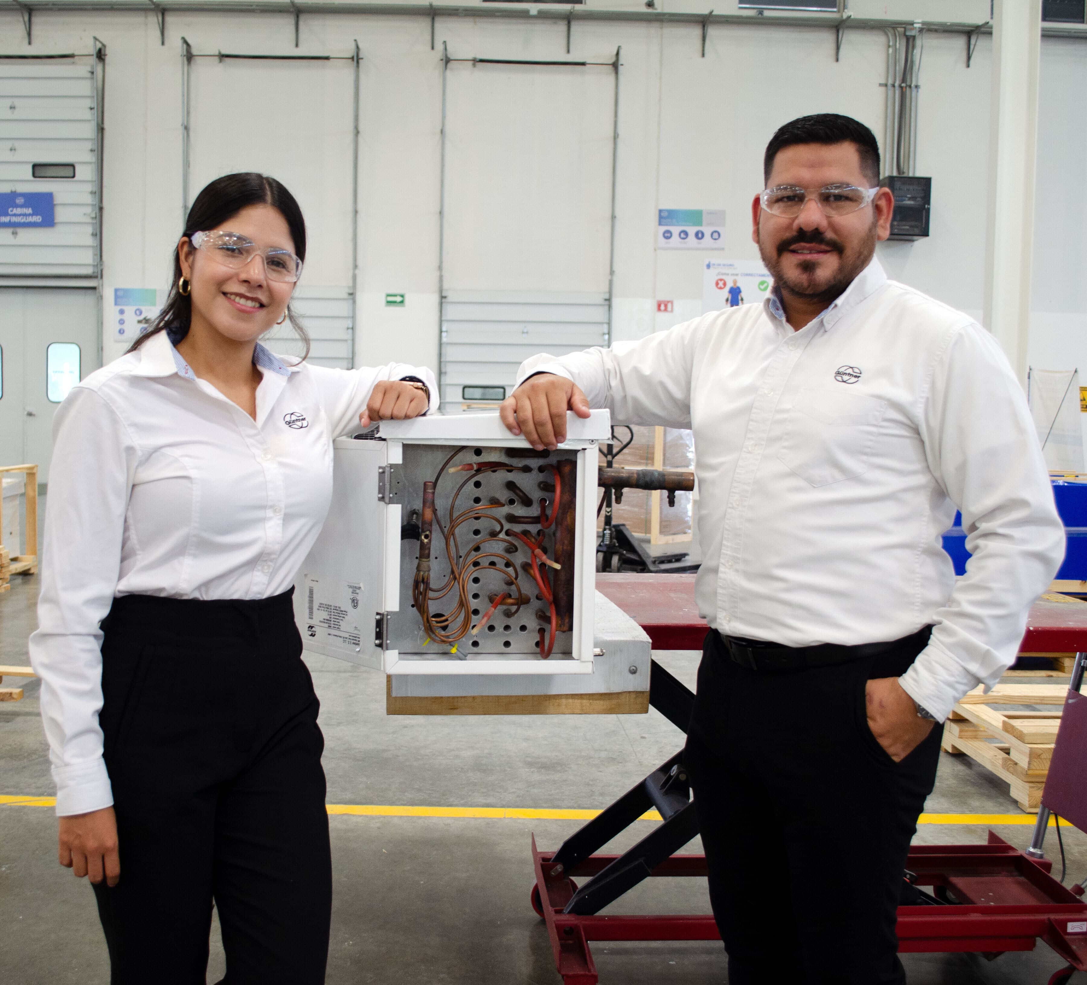 Two people in safety glasses and white shirts stand beside an open electrical cabinet, showing copper wires, in a factory workshop.