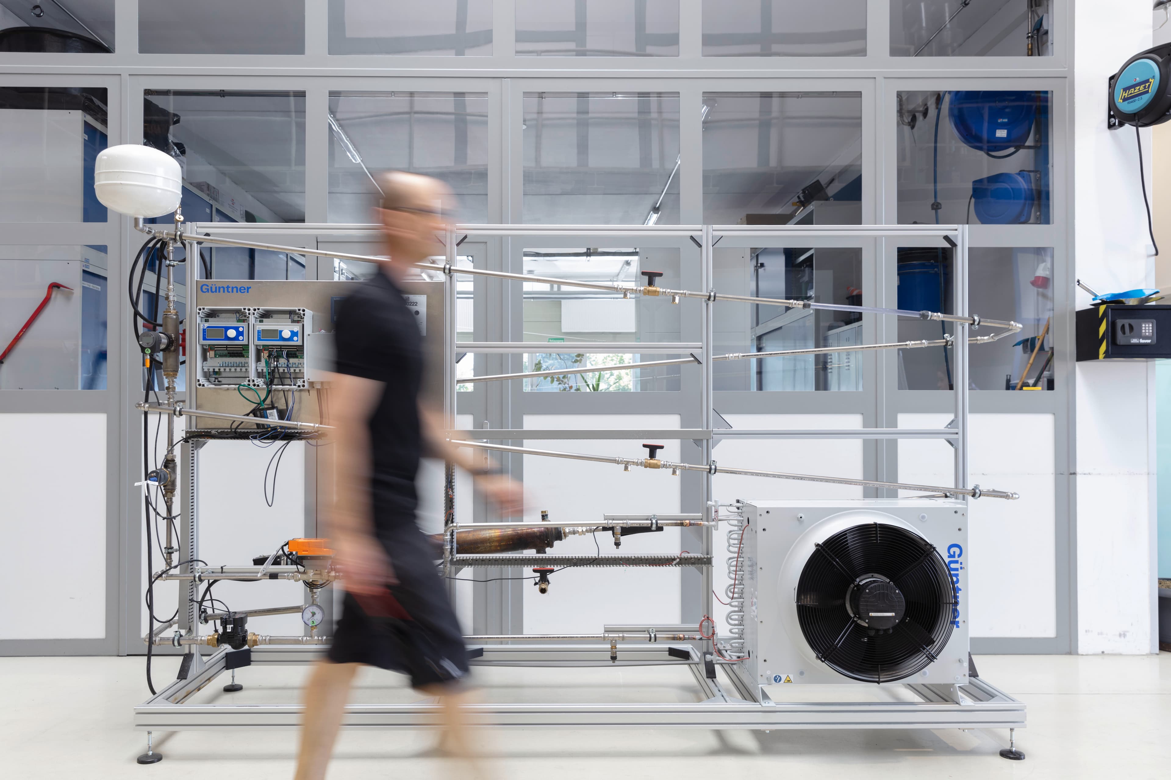 Blurred person walks past a metal lab rig with tubes, gauges and controls; glass walls behind and a large cooling unit with a fan on the right.