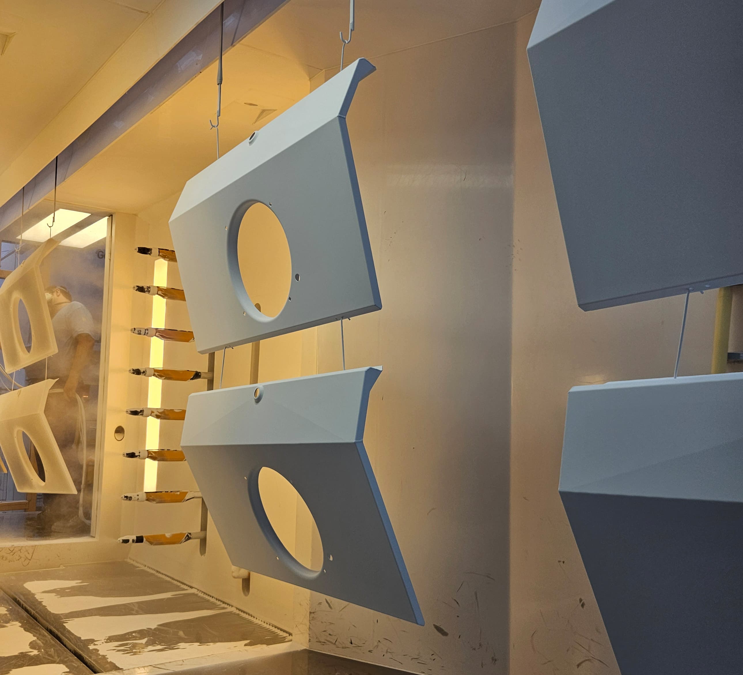 Industrial paint booth with pale blue, faceted metal panels hanging on wires; circular cutouts in panels and a person visible behind a glass wall.