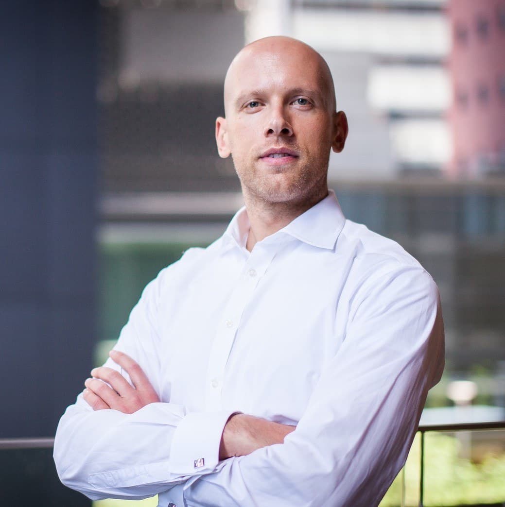 Bald person in a white button-up shirt stands with arms crossed, facing the camera against a blurred urban backdrop.